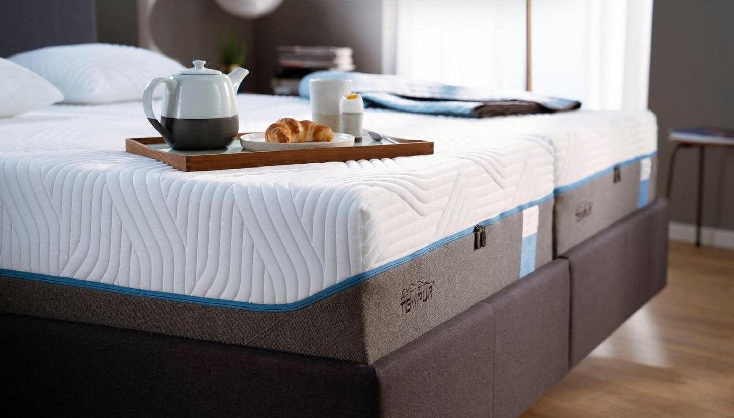 Hotelowe łóżko kontynentalne, na którym znajduje się materac. Na materacu umieszczono tacę z kawą, herbatą oraz rogalikiem.